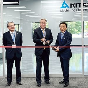 Three men in formal attire participate in a ribbon-cutting ceremony at the new Kitamura U.S. corporate headquarters, with the Kitamura logo visible on the glass wall behind them.