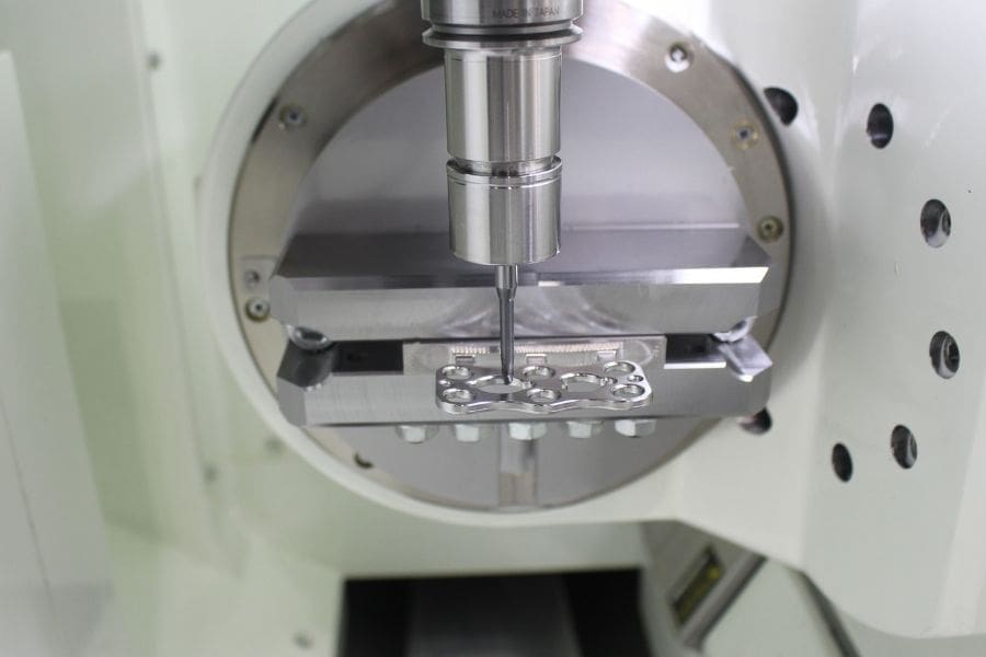 Close-up of a 5-axis CNC machine performing precision milling on a small, complex metal component held in a custom fixture on a rotary table.